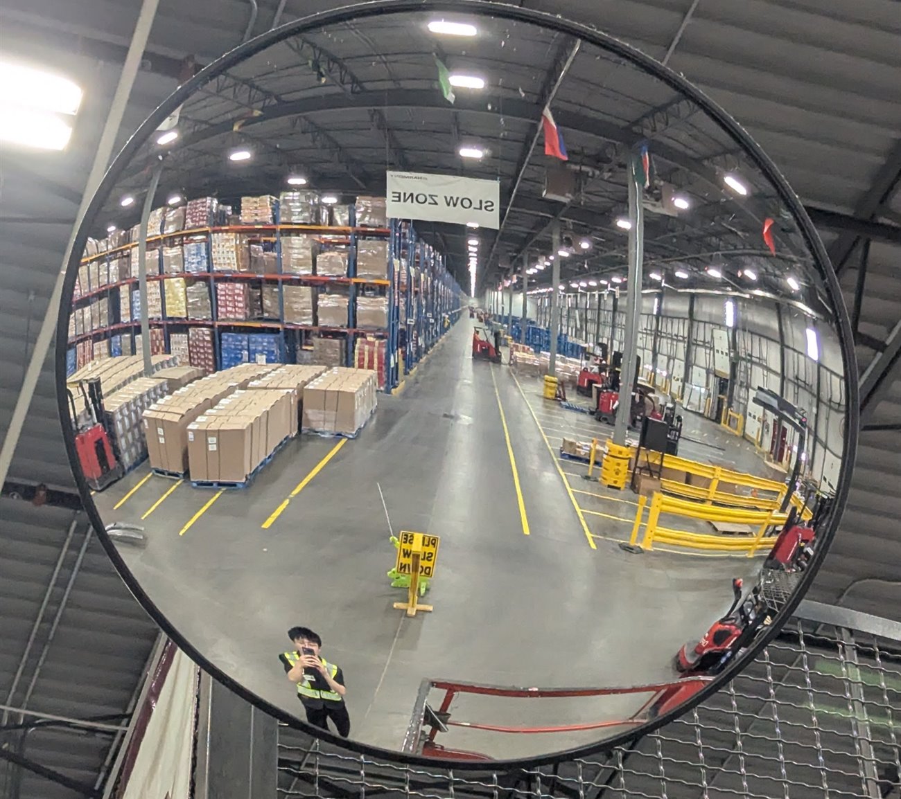 A convex safety mirror reflecting a DHL warehouse aisle lined with racking and pallets